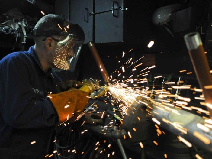 A person wearing protective gear, including a face shield and gloves, is using a power tool that produces sparks while working on a metal object. The background is dimly lit, highlighting the bright sparks flying from the tool. Various tools and equipment are visible on the workbench, suggesting a workshop environment.