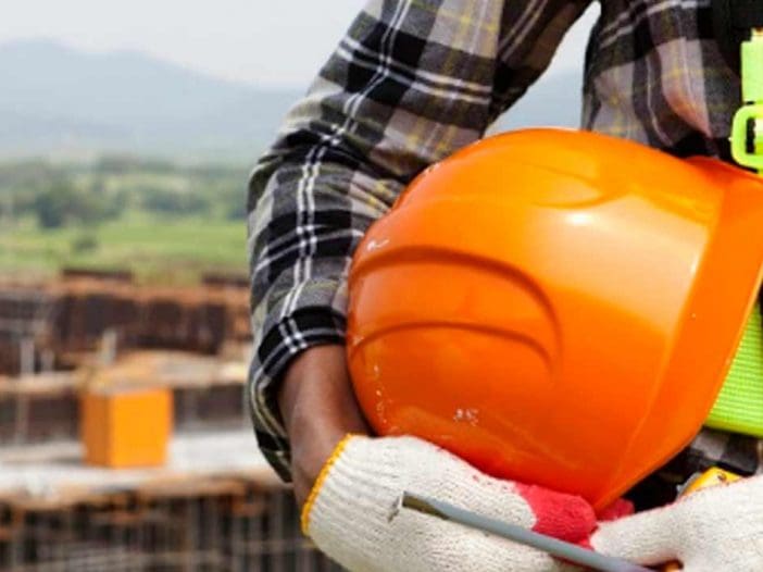 A construction worker is holding an orange hard hat under their arm. They are wearing a plaid shirt and a safety harness with bright green straps. The worker's hands are protected by white gloves with red and yellow accents. In the background, a construction site is visible, featuring scaffolding and a crane, with green hills in the distance.