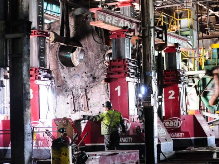A large industrial setting features heavy machinery, specifically two hydraulic jacks labeled "1" and "2," positioned to lift a massive, metallic object with a circular opening. A worker in a reflective yellow vest is interacting with the equipment, while various tools and equipment are visible in the background, including a gas cylinder and scaffolding. The environment is dimly lit, with bright lights illuminating the machinery and the worker.
