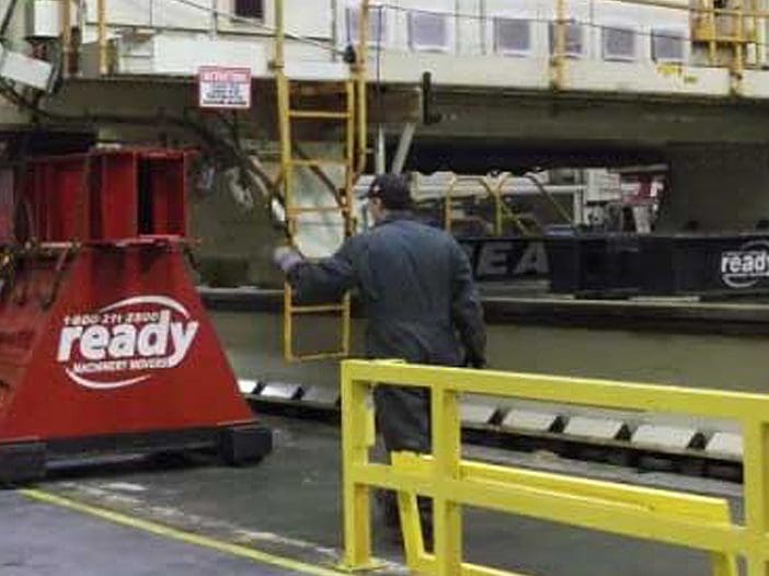 A warehouse or industrial setting is depicted, featuring large machinery and equipment. In the foreground, a red forklift is positioned next to a prominent red and white block labeled "ready." A worker in a dark jumpsuit is seen gesturing towards the machinery above, which is mounted on a raised platform. The environment is well-lit, with yellow safety barriers and various signs visible in the background, indicating a busy operational area.
