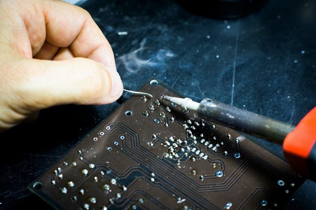 A hand is holding a thin wire while a soldering iron is being used to solder it onto a printed circuit board (PCB). The PCB has various metallic components and traces visible, with some smoke rising from the soldering process. The background is a dark surface, enhancing the focus on the soldering activity.