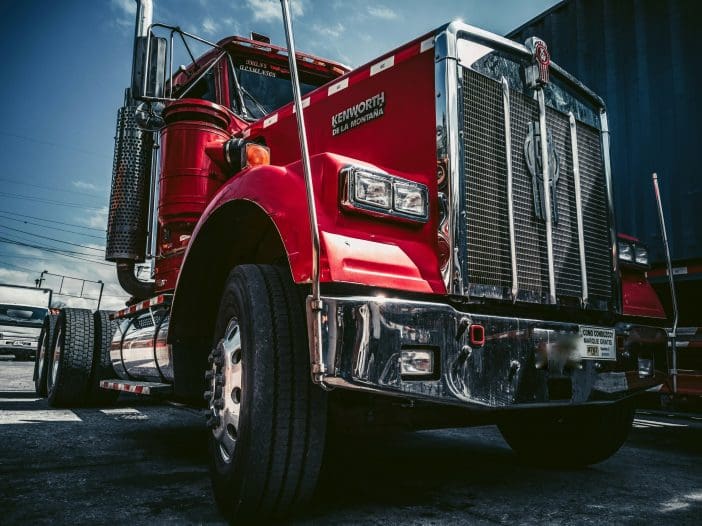 A large red semi-truck is prominently featured, showcasing its shiny chrome grille and distinctive Kenworth branding. The truck has a polished appearance with a tall exhaust stack and large tires. The background includes a hint of other vehicles and a cloudy sky, adding depth to the scene. The angle emphasizes the truck's robust build and striking color.