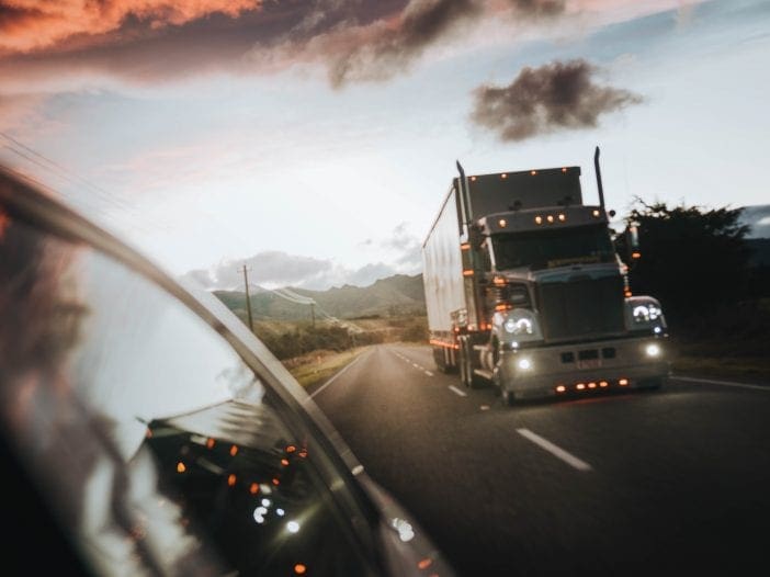 A large semi-truck is driving on a two-lane road, captured from a side view. The truck has a shiny silver exterior with illuminated lights, and it is positioned in the right lane. In the foreground, the reflection of the truck can be seen in the side mirror of a car. The background features a scenic landscape with rolling hills and a colorful sky at dusk, filled with clouds and warm hues of orange and pink.