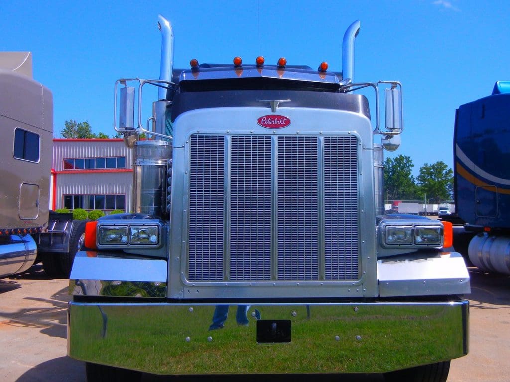 A shiny, chrome-fronted truck is prominently displayed, showcasing its large grille and dual headlights. The truck features a distinctive design with a tall exhaust stack and orange clearance lights on the roof. In the background, a building with large windows and red accents is visible, along with another truck partially obscured on the right. The sky is clear and blue, adding to the bright, sunny atmosphere.