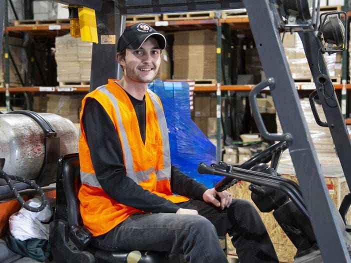 A young man sits in a forklift, wearing an orange safety vest over a black long-sleeve shirt and a cap. He is smiling and appears to be in a warehouse setting, with shelves stocked with boxes in the background. The forklift has a gas tank visible on the side, and there are blue plastic wraps and wooden crates nearby. The environment is well-lit and organized.