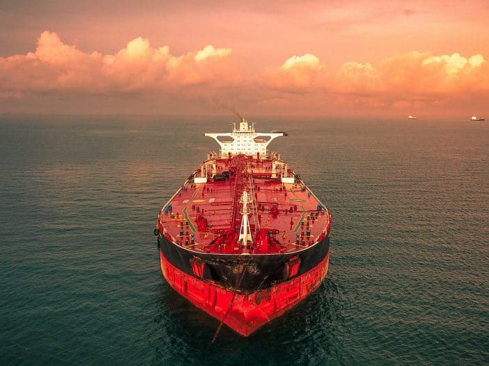 A large oil tanker is depicted, viewed from the front, cutting through calm waters. The ship's hull is predominantly red with a black bottom, and it features various structures on the deck, including loading equipment and safety railings. The background showcases a serene ocean under a sky filled with soft, orange-hued clouds, suggesting either sunrise or sunset. Additional ships can be seen in the distance, adding to the maritime scene.