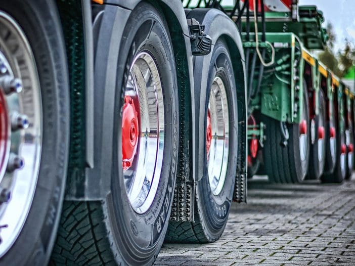 A row of large truck tires is prominently displayed, showcasing their robust design. The tires have deep treads for traction, and the rims are shiny with a red center cap. The perspective emphasizes the size and alignment of the tires, which are part of a heavy-duty vehicle. The background features a blurred view of additional trucks, suggesting a commercial or industrial setting. The ground is paved with square stones, adding texture to the scene.