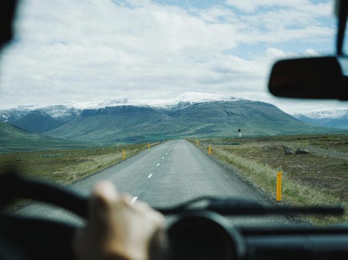 A winding road stretches into the distance, flanked by green fields and rocky terrain. Snow-capped mountains rise majestically in the background under a partly cloudy sky. The view is from inside a vehicle, with a hand gripping the steering wheel visible in the foreground. Yellow road markers line the sides of the road, guiding the way through the scenic landscape.