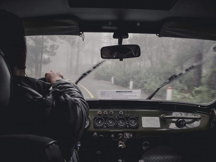 A view from inside a vehicle shows a person driving on a winding road surrounded by fog. The driver's hand is on the steering wheel, and the dashboard features various gauges and controls. Rain is visible on the windshield, with wipers in motion. The scenery outside includes blurred outlines of trees and road markers, emphasizing the misty atmosphere.