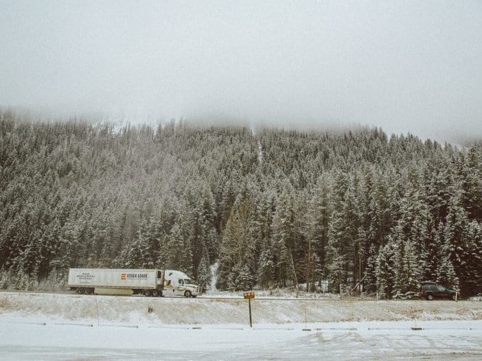 A large white truck with a trailer is driving along a snowy road, flanked by dense evergreen trees dusted with snow. The background features a misty mountain range partially obscured by clouds. A dark-colored vehicle is parked on the roadside, and the ground is covered in a layer of snow, creating a serene winter landscape.