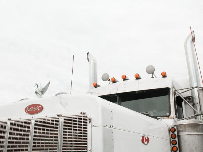 A white Peterbilt truck is prominently featured, showcasing its distinctive front grille with horizontal slats. Atop the cab, a chrome ornament in the shape of a bird with outstretched wings is visible. The truck has several orange lights along the roof and two tall exhaust pipes on either side. The background features a cloudy sky, adding a muted tone to the scene.