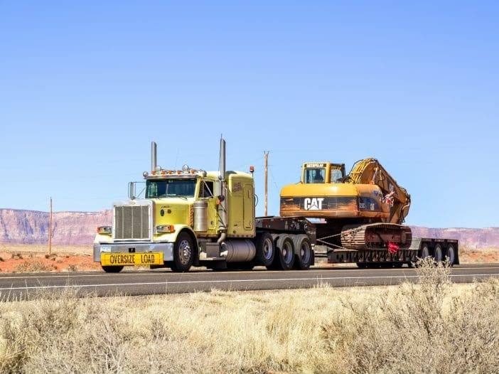 A large yellow semi-truck is transporting an oversized Caterpillar excavator on a flatbed trailer. The truck features a prominent "OVERSIZE LOAD" sign on the front. The background showcases a clear blue sky and a desert landscape with distant red rock formations. Sparse vegetation is visible along the roadside.