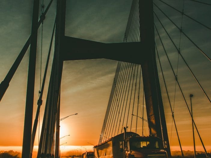A large truck is driving on a bridge during sunset, with the sun low on the horizon casting a warm glow. The bridge features tall, angular supports and cables, creating a striking silhouette against the colorful sky. The road is illuminated by streetlights, and the scene conveys a sense of tranquility and movement.