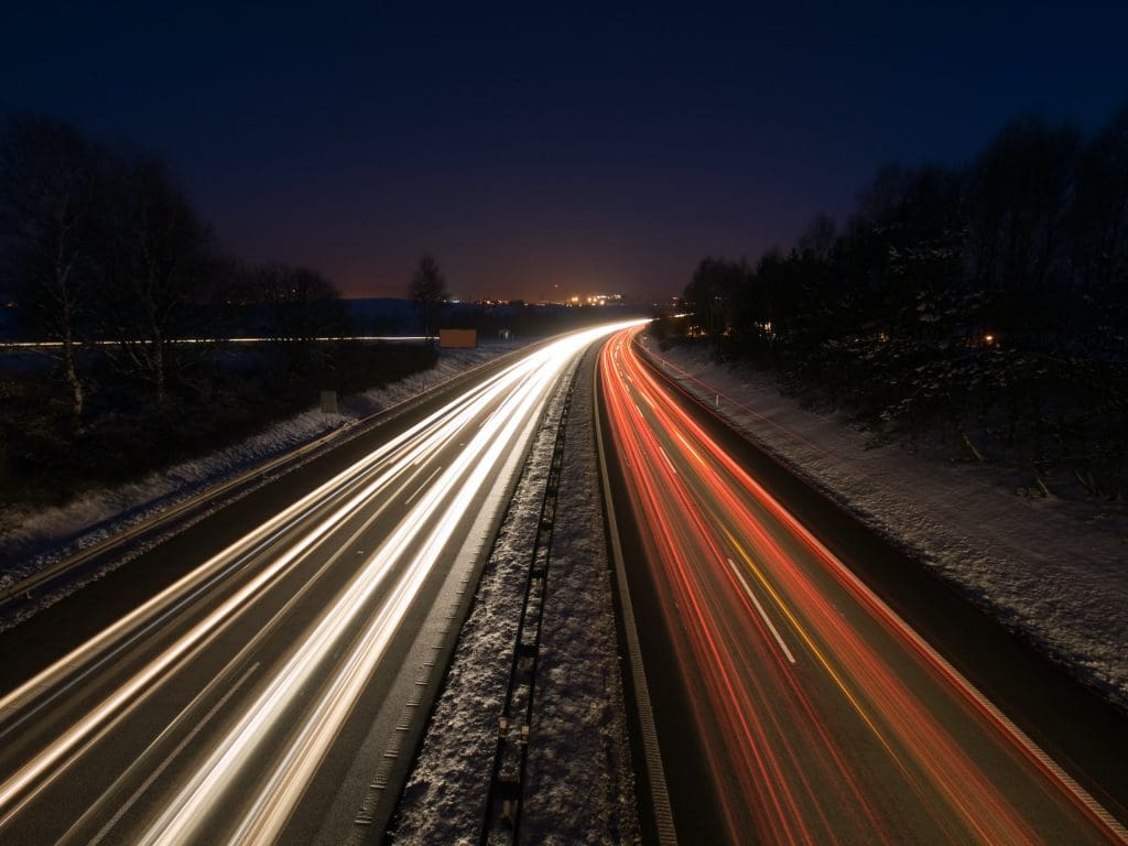 A nighttime highway scene features streaks of light from moving vehicles, creating a dynamic effect. The road is flanked by snow-covered ground and trees, with faint lights from distant buildings visible in the background. The contrast between the dark sky and the illuminated road enhances the sense of motion and depth in the image.