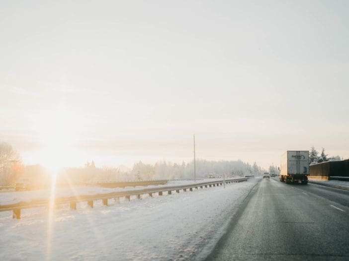 A snow-covered highway stretches into the distance, flanked by a guardrail on the left. The scene is bathed in soft, golden light from a low sun, creating a warm glow against the cool tones of the snow. A large truck is seen traveling on the right side of the road, while a smaller vehicle is visible in the distance. Trees dusted with snow line the horizon, contributing to the serene winter landscape.