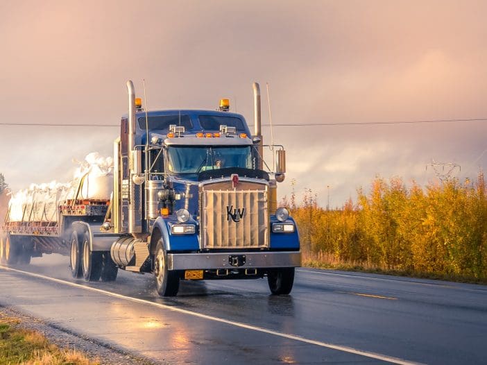 A large blue semi-truck is driving along a wet road, with a trailer carrying white cylindrical objects. The truck features a prominent front grille with the letters "KW" and has multiple chrome exhaust stacks. Surrounding the road are trees with autumn foliage, and the sky is overcast, creating a misty atmosphere. The scene conveys a sense of movement and transportation in a rural setting.
