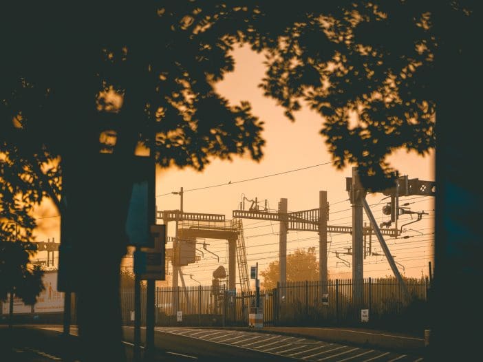 Silhouetted against a warm, golden sky, a railway scene features overhead power lines and support structures. The foreground includes tree branches and a bus stop sign, while a fence lines the railway area. The atmosphere suggests early morning or late evening, with soft lighting enhancing the tranquil setting.