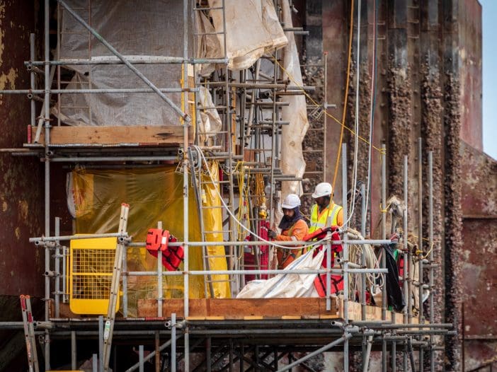 Two construction workers are positioned on a scaffold platform, working on a large structure. They are wearing hard hats and safety vests. The scaffold is made of metal and wood, with safety nets and tarps partially covering the area. In the background, the weathered surface of the structure shows signs of wear, with peeling paint and barnacles visible. Tools and equipment are scattered around the platform, indicating ongoing maintenance or repair work.