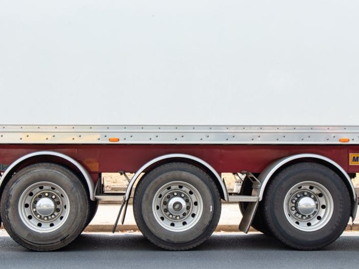 A side view of a truck trailer shows three large wheels with metallic rims. The trailer has a white exterior with a red lower section. The wheels are mounted on a silver chassis, and the tires appear robust, suitable for heavy loads. The ground is asphalt, and there is a slight shadow cast by the trailer.
