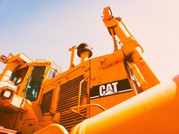 A large yellow bulldozer is prominently displayed, showcasing its cab and machinery. The vehicle features a visible logo reading "CAT" on its side. The image is taken from a low angle, emphasizing the height and structure of the bulldozer against a clear blue sky. The sunlight creates a warm tone, highlighting the details of the machine's design and components.