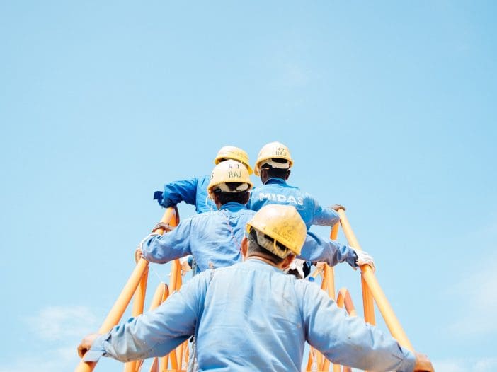 A group of construction workers is climbing a ladder, ascending towards a bright blue sky. They are wearing yellow hard hats and blue work shirts. The focus is on their backs as they grasp the ladder's orange rungs, showcasing teamwork and determination in a construction setting.