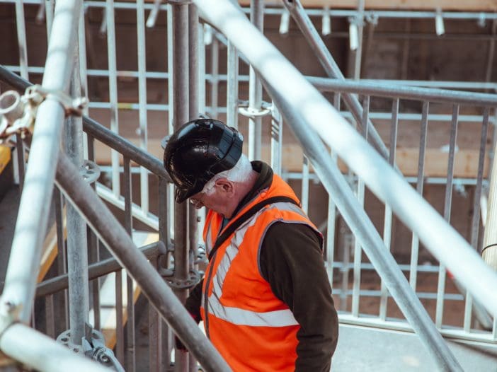 A construction worker is wearing a black hard hat and an orange safety vest with reflective stripes. He is standing on a staircase surrounded by scaffolding, looking down at the steps. The background features wooden planks and metal supports typical of a construction site. The worker appears focused on his task.
