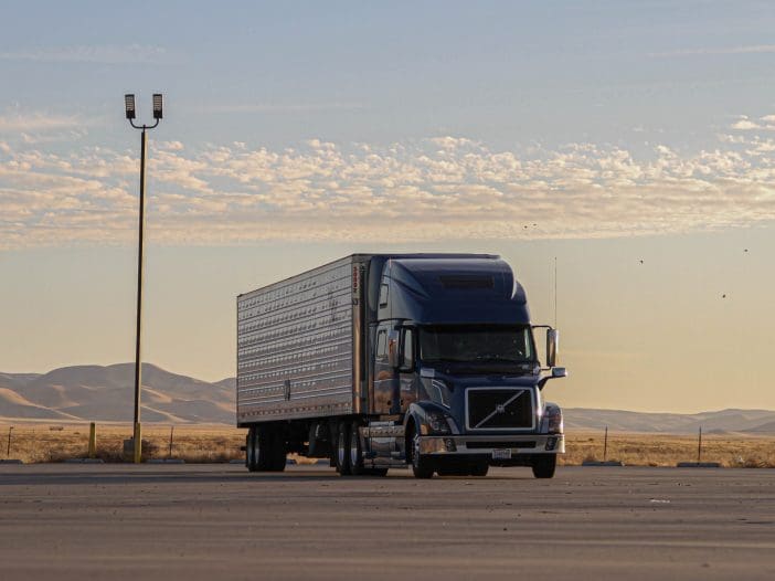 A blue semi-truck is parked on a wide, empty lot with a smooth asphalt surface. The truck has a silver trailer with a corrugated exterior. In the background, rolling hills are visible under a sky filled with soft, wispy clouds. A tall light pole stands nearby, casting a shadow on the ground. The scene is bathed in warm, golden light, suggesting early morning or late afternoon.