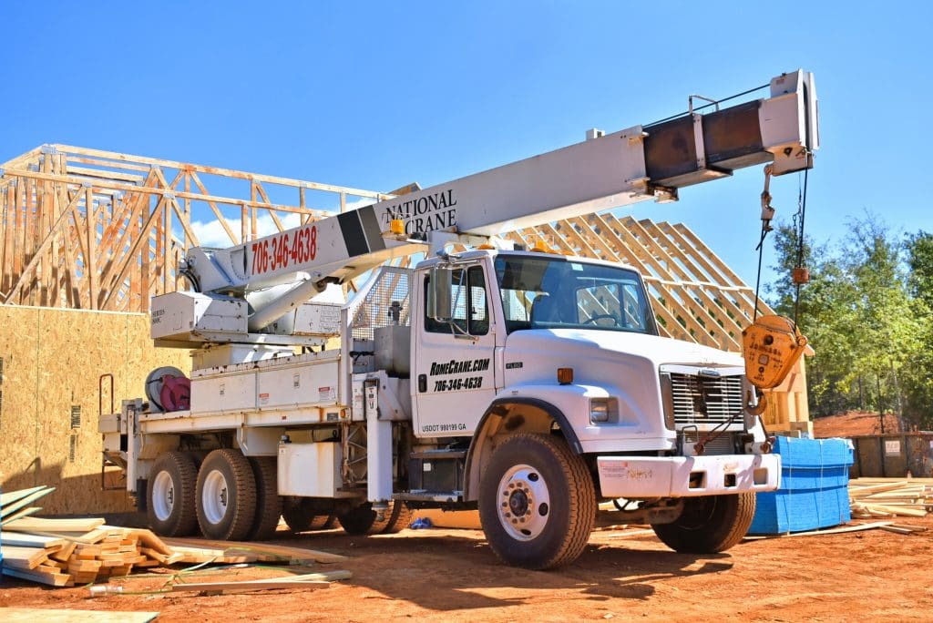 A large white crane truck is positioned on a construction site, with a boom extended upward. The truck features a flatbed with a winch and is equipped for lifting heavy materials. In the background, a wooden frame of a building is partially constructed, showcasing exposed beams. Nearby, stacks of lumber and blue tarps are visible on the ground, indicating ongoing construction activity. The sky is clear and blue, suggesting a sunny day.