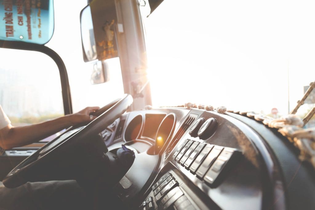 The interior of a bus is shown, focusing on the driver's seat and dashboard. A hand is gripping the steering wheel, while various controls and buttons are visible on the dashboard. The sunlight is streaming in through the windshield, creating a warm glow. The scene conveys a sense of motion and readiness for travel.