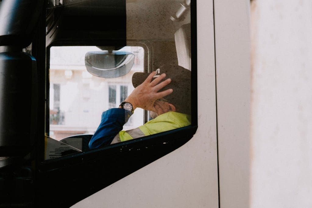 A person is seated inside a truck, visible through the driver's side window. They are wearing a blue jacket and a bright yellow safety vest. The individual is holding their hand to their head, possibly in thought or frustration. A watch is visible on their wrist. The background shows a blurred view of buildings outside the truck. The window appears slightly dirty, adding a textured look to the image.