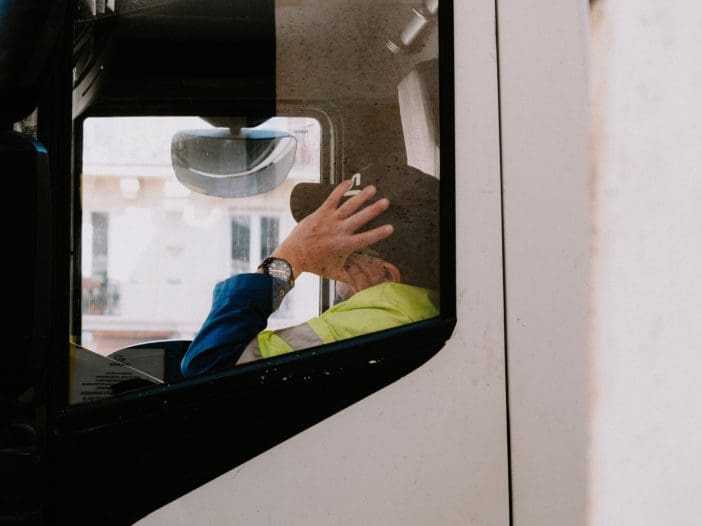 A person is seated inside a truck, visible through the driver's side window. They are wearing a blue jacket and a bright yellow safety vest. The individual is holding their hand to their head, possibly in thought or frustration. A watch is visible on their wrist. The background shows a blurred view of buildings outside the truck. The window appears slightly dirty, adding a textured look to the image.