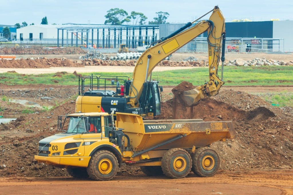 A yellow Volvo A30F articulated dump truck is positioned on a construction site, with its bed raised to receive dirt from a nearby excavator. The excavator, also yellow and branded with CAT, is actively digging into a mound of earth. In the background, various construction materials and equipment are visible, along with partially constructed buildings and green grass areas. The scene conveys a busy construction environment.