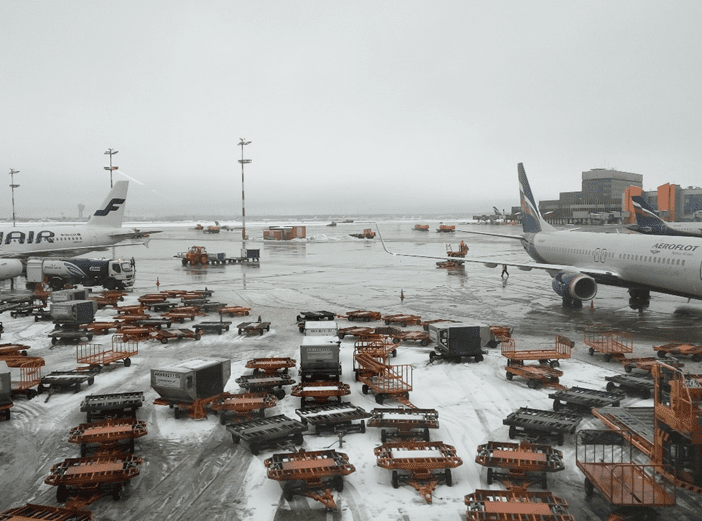 An airport scene features two airplanes parked on a snowy tarmac. On the left, a Finnair aircraft is visible, while on the right, an Aeroflot plane is stationed. The ground is cluttered with luggage carts and containers, some of which are orange and black. The sky is overcast, contributing to a gray atmosphere, and various airport structures can be seen in the background.