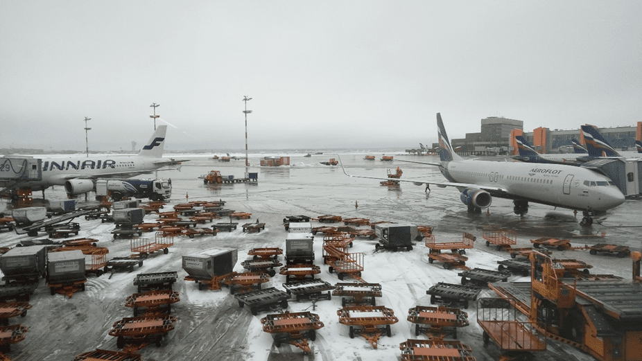 An airport scene features two airplanes parked on a snowy tarmac. On the left, a Finnair aircraft is visible, while on the right, an Aeroflot plane is stationed. The ground is cluttered with luggage carts and containers, some of which are orange and black. The sky is overcast, contributing to a gray atmosphere, and various airport structures can be seen in the background.