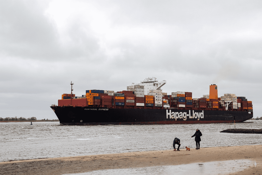 A large cargo ship, identified as "Guayaquil Express," is navigating through a waterway. The ship is heavily loaded with colorful shipping containers stacked on its deck, prominently displaying the Hapag-Lloyd logo. In the foreground, two people are on the sandy shore, one crouching down and the other standing, with a small dog nearby. The sky is overcast, creating a moody atmosphere.