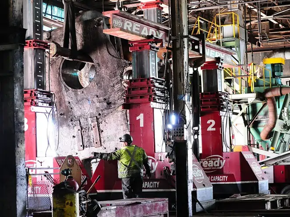 A large industrial setting features two hydraulic presses, labeled "1" and "2," positioned on either side of a massive metal component being lifted. The presses are red with visible branding and are equipped with hydraulic cylinders. A worker in a green safety jacket and helmet is operating the machinery, focusing on the lifting process. The background includes various industrial equipment and structures, with overhead lighting illuminating the scene. The atmosphere is busy and functional, typical of a manufacturing or repair facility.