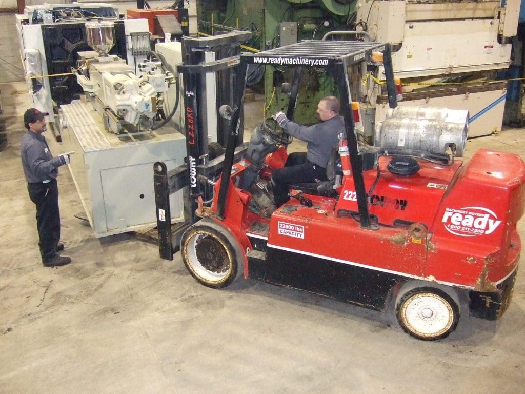 A red forklift is positioned in a warehouse, lifting a large piece of machinery from a gray platform. The forklift operator, wearing gloves and glasses, is focused on maneuvering the load. Nearby, a worker in a gray uniform is pointing towards the machinery, possibly giving instructions. The warehouse has various industrial equipment in the background, with concrete flooring and overhead lighting.