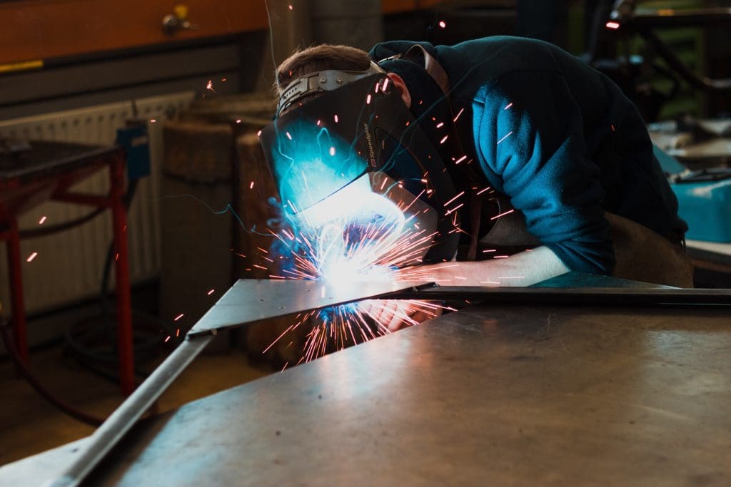 A person is welding metal pieces together in a workshop. They are wearing protective gear, including a welding helmet, and are focused on the task. Bright sparks are flying from the welding area, illuminating the workspace. The background features various tools and equipment typical of a metalworking environment.