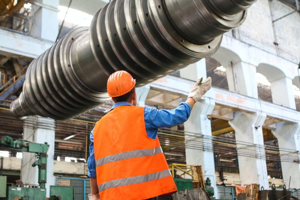 A worker in an orange safety vest and hard hat stands in a large industrial facility, looking up at a massive metallic cylindrical object being suspended above. The worker is gesturing with one hand, while wearing gloves on both hands. The background features machinery and equipment typical of a manufacturing environment, with high ceilings and large windows allowing natural light to enter.