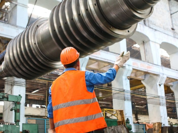 A worker in an orange safety vest and hard hat stands in a large industrial facility, looking up at a massive metallic cylindrical object being suspended above. The worker is gesturing with one hand, while wearing gloves on both hands. The background features machinery and equipment typical of a manufacturing environment, with high ceilings and large windows allowing natural light to enter.