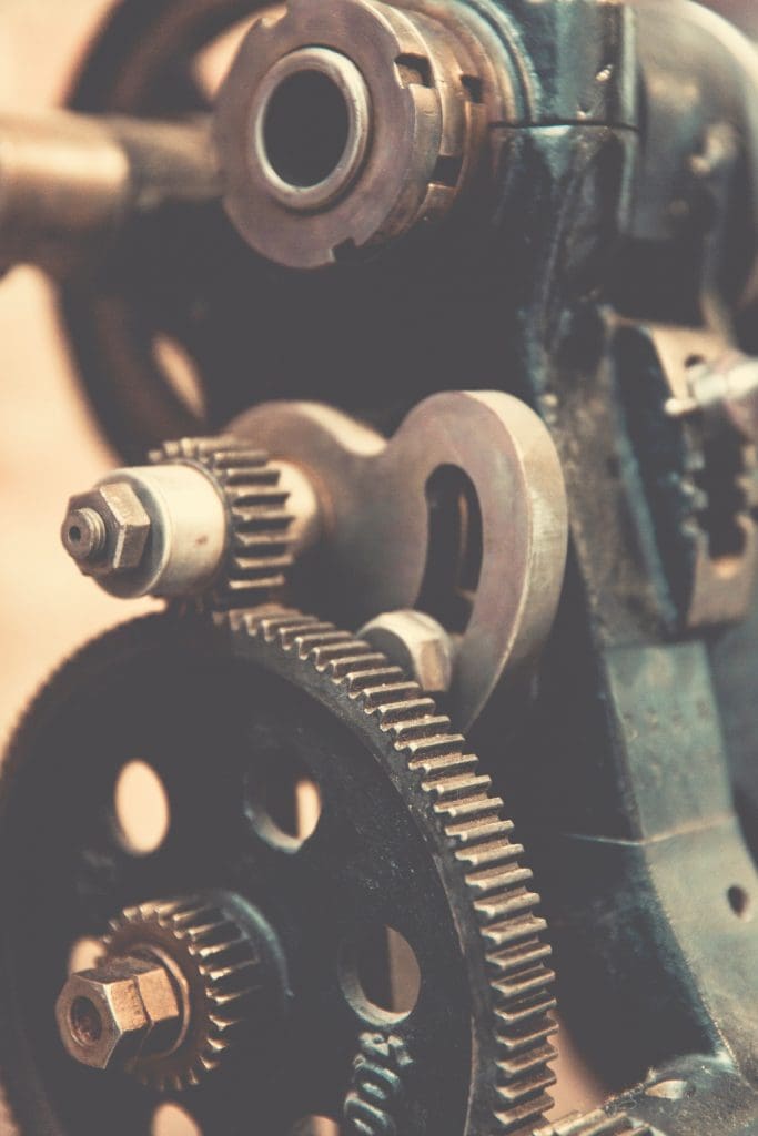 A close-up view of a mechanical assembly featuring several gears and metal components. The image highlights a large black gear with holes and smaller metallic gears attached to it. There are also bolts and a cylindrical part, suggesting a complex machinery setup. The overall aesthetic is vintage, with a slightly worn appearance.