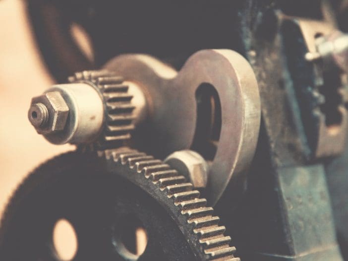 A close-up view of a mechanical assembly featuring several gears and metal components. The image highlights a large black gear with holes and smaller metallic gears attached to it. There are also bolts and a cylindrical part, suggesting a complex machinery setup. The overall aesthetic is vintage, with a slightly worn appearance.
