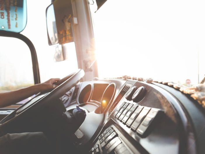 The interior of a bus is visible, focusing on the driver's seat and dashboard. A hand is gripping the steering wheel, while the dashboard features various buttons and controls. Sunlight is streaming in from the front, creating a warm glow. The scene suggests an active driving environment, with a hint of dust on the dashboard, indicating use over time.