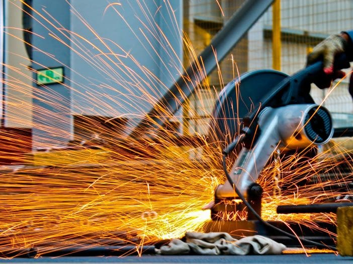 A cutting tool is in use, generating a burst of bright orange sparks as it makes contact with metal. The tool is positioned on a surface with a cloth nearby, and the background features industrial elements, including a blurred structure and safety signage. The scene conveys a dynamic and energetic atmosphere typical of metalworking or fabrication activities.