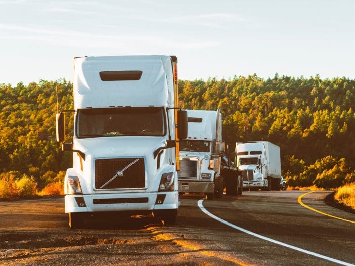 A line of three white semi-trucks is parked along a winding road. The first truck, prominently displayed, has a distinctive Volvo front grille. The background features lush green hills illuminated by warm sunlight, creating a serene atmosphere. The road curves gently, leading into the distance.