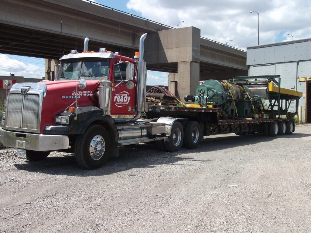 A red and silver semi-truck is parked on a gravel surface, featuring a prominent front grille and tall exhaust stacks. The truck is towing a flatbed trailer that carries large industrial machinery, including a green cylindrical component and additional equipment secured with straps. The background includes a concrete overpass and a building, with a partly cloudy sky overhead.