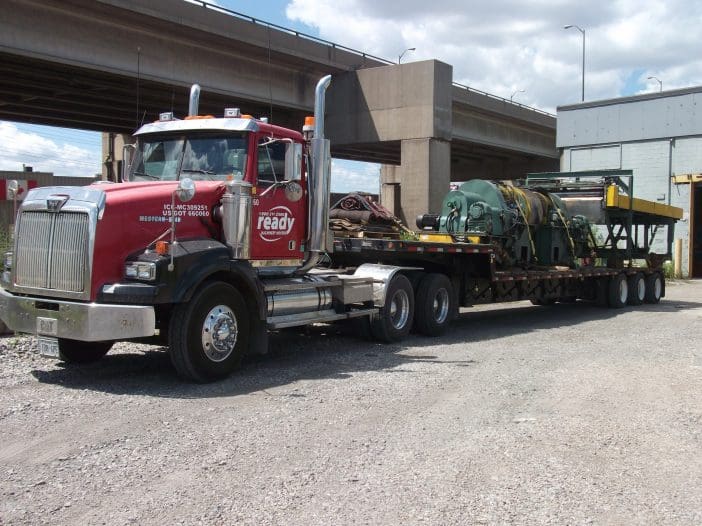 A red and silver semi-truck is parked on a gravel surface, with a long flatbed trailer attached. The truck features a prominent front grille and large exhaust stacks. On the trailer, there is heavy machinery, including a large green piece of equipment secured with straps. The background shows a concrete overpass and a building with a gray exterior. The sky is partly cloudy.