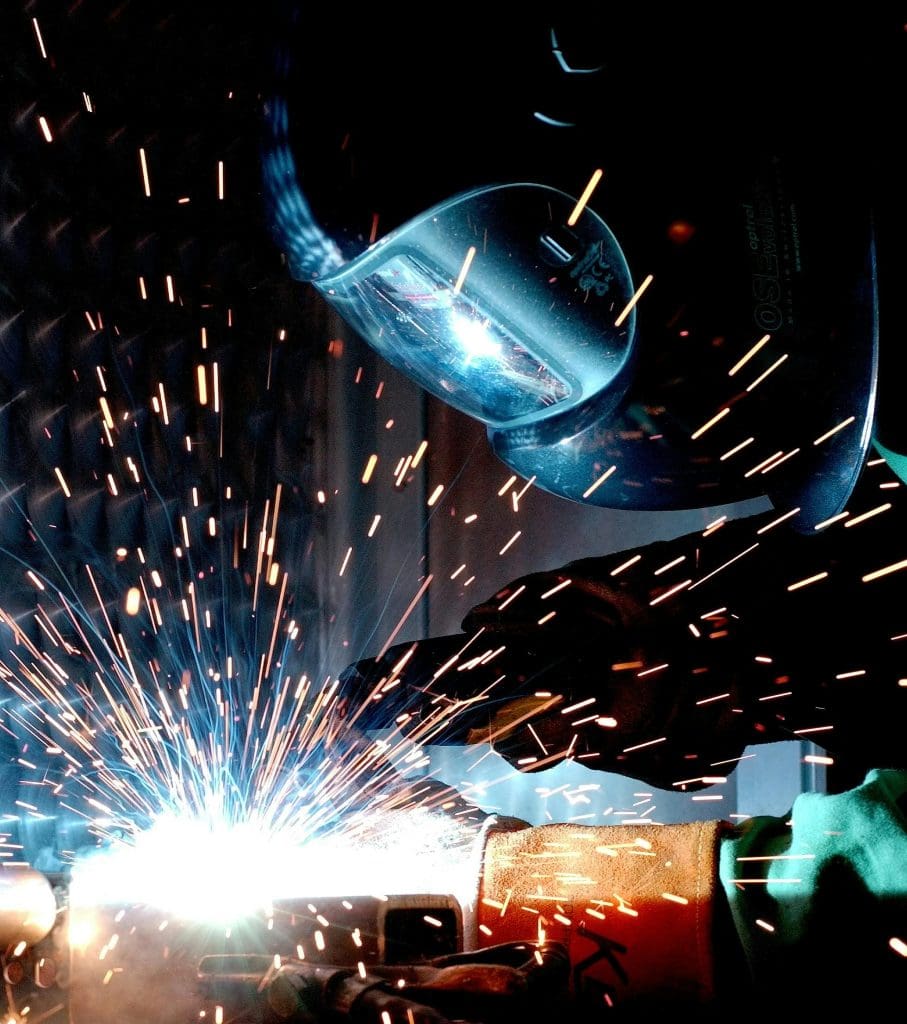A welder is engaged in the process of welding, with bright sparks flying in various directions. The welder is wearing protective gear, including a helmet with a clear visor and gloves. The focus is on the welding torch, which emits a bright blue-white light as it makes contact with the metal. The background is dark, highlighting the vivid sparks and the intensity of the welding operation.