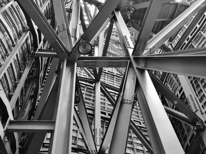 A complex arrangement of steel beams and structural supports is visible, showcasing an intricate lattice of intersecting angles and lines. The image is in black and white, emphasizing the geometric patterns and textures of the metal. Above, a grid-like structure can be seen, suggesting an open space or roof. The overall composition creates a sense of depth and perspective, drawing the viewer's eye upward through the framework.