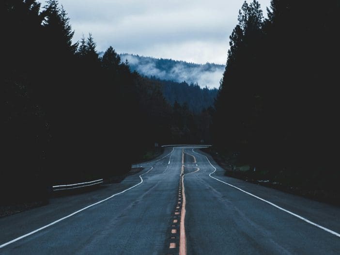 A winding road stretches into the distance, flanked by dense evergreen trees on both sides. The asphalt is dark and wet, reflecting the overcast sky above. A faint orange line runs down the center, guiding the viewer's eye toward the horizon where misty mountains are partially obscured by clouds. The overall atmosphere is moody and tranquil, with a sense of solitude.